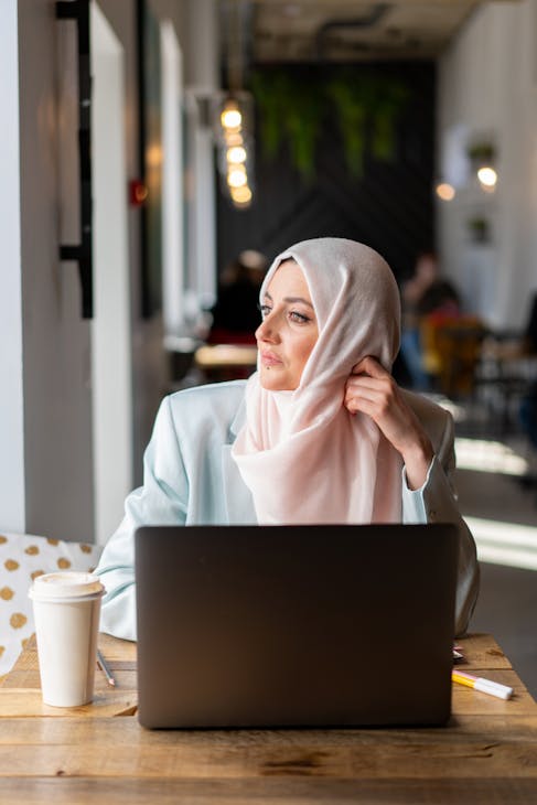 A woman in traditional attire thoughtfully working on a laptop in a cozy coffee shop.